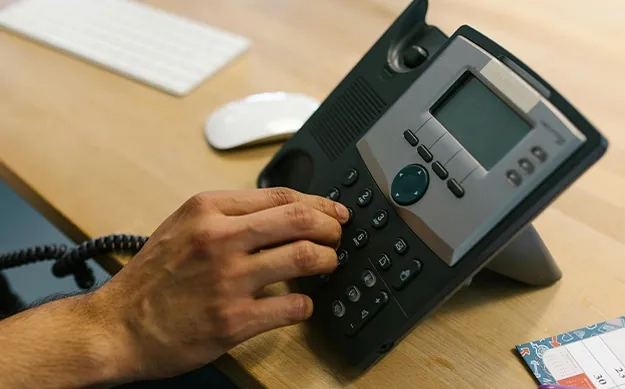 Une main composant un numéro sur le clavier d'un téléphone de bureau IP noir, posé sur un bureau en bois à côté d'une souris et d'un clavier d'ordinateur.