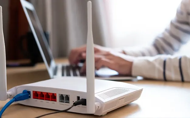 Gros plan sur un routeur Wi-Fi blanc avec deux antennes sur un bureau, tandis qu'une personne travaille sur un ordinateur portable flou en arrière-plan.