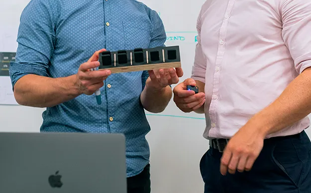 Deux professionnels en pleine séance de collaboration sur un projet d'outsourcing IT, examinant un prototype physique devant un ordinateur portable et un tableau blanc de brainstorming.